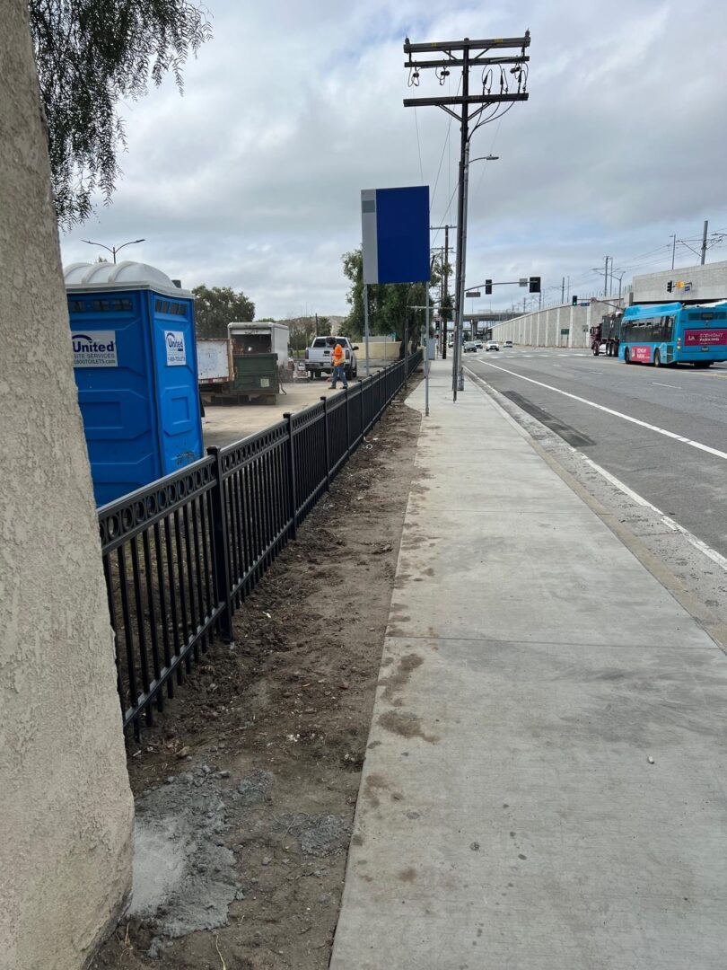 Sidewalk with a fence and a blue porta-potty.