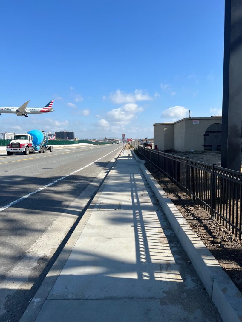 Sidewalk and road with plane overhead.