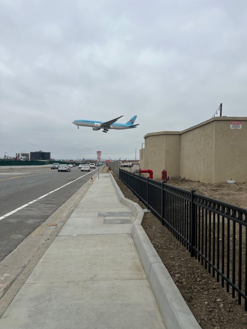 Airplane landing over a sidewalk and fence.